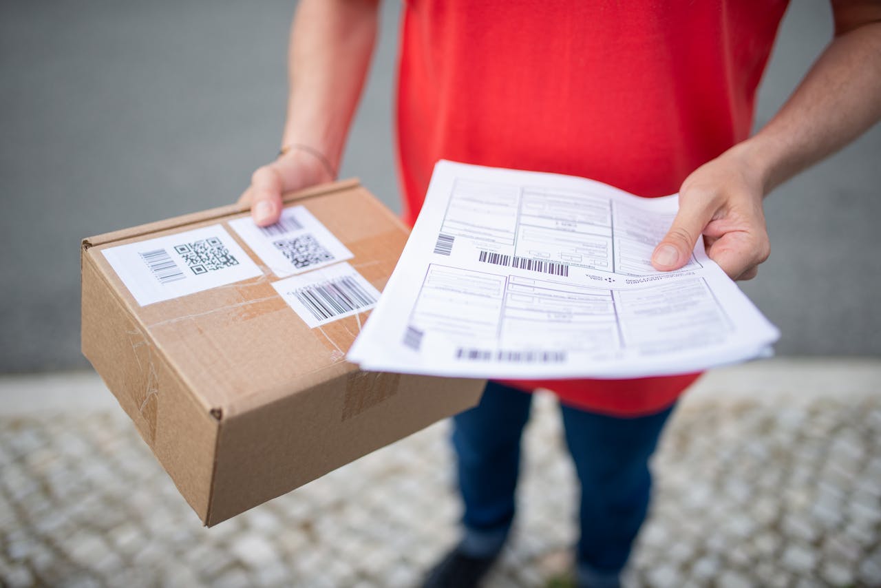 about-img Close-up of person in red shirt holding a brown package and delivery documents outside.
