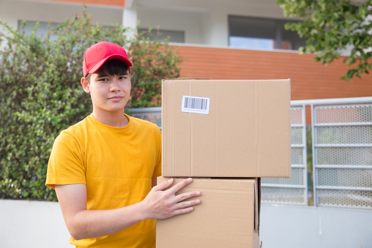 gallery-4 Asian delivery man holding cardboard boxes in an outdoor setting, wearing a red cap and yellow shirt.